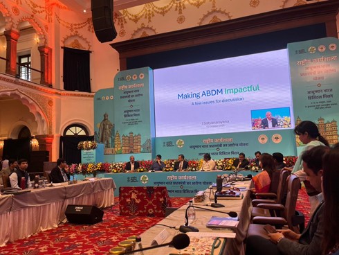 A grand conference hall with ornate walls and red carpet features a long table with multiple panelists including men and women in formal attire seated and speaking into microphones surrounded by floral arrangements and documents. A large screen displays Making ABDM Impactful in English and Hindi with logos of government health initiatives and images of participants. The stage has decorative backdrops with cityscape motifs and event branding in blue and white. Additional screens show related visuals and the audience area is visible in the background.