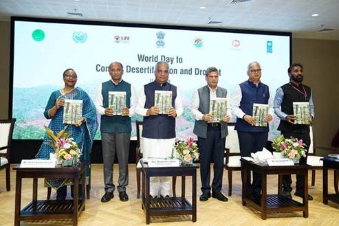 The image shows a group of people, presumably dignitaries, standing in a room with decorations and documents related to environmental and lifestyle themes, including flags and titles of individuals.