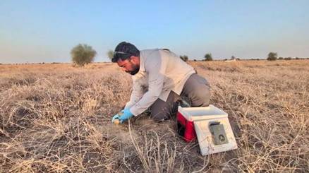 A man in a white shirt and khaki pants kneels on dry grass, examining a weather instrument under a clear sky.AI-generated content may be incorrect.