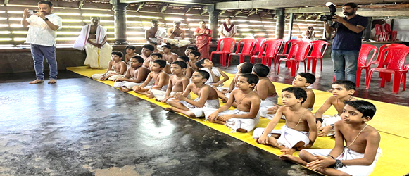 A group of boys sitting on yellow mats in a room with a person standing in front of themAI-generated content may be incorrect.