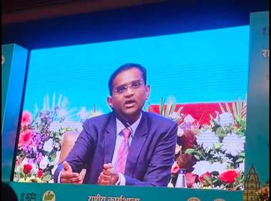 A man in a dark suit and red tie with glasses speaks on a large blue screen displaying Hindi text about Rashtriya Karyashala and Ayushman Bharat programs surrounded by floral decorations and event logos including ABDM and NHA emblems in a formal setting.